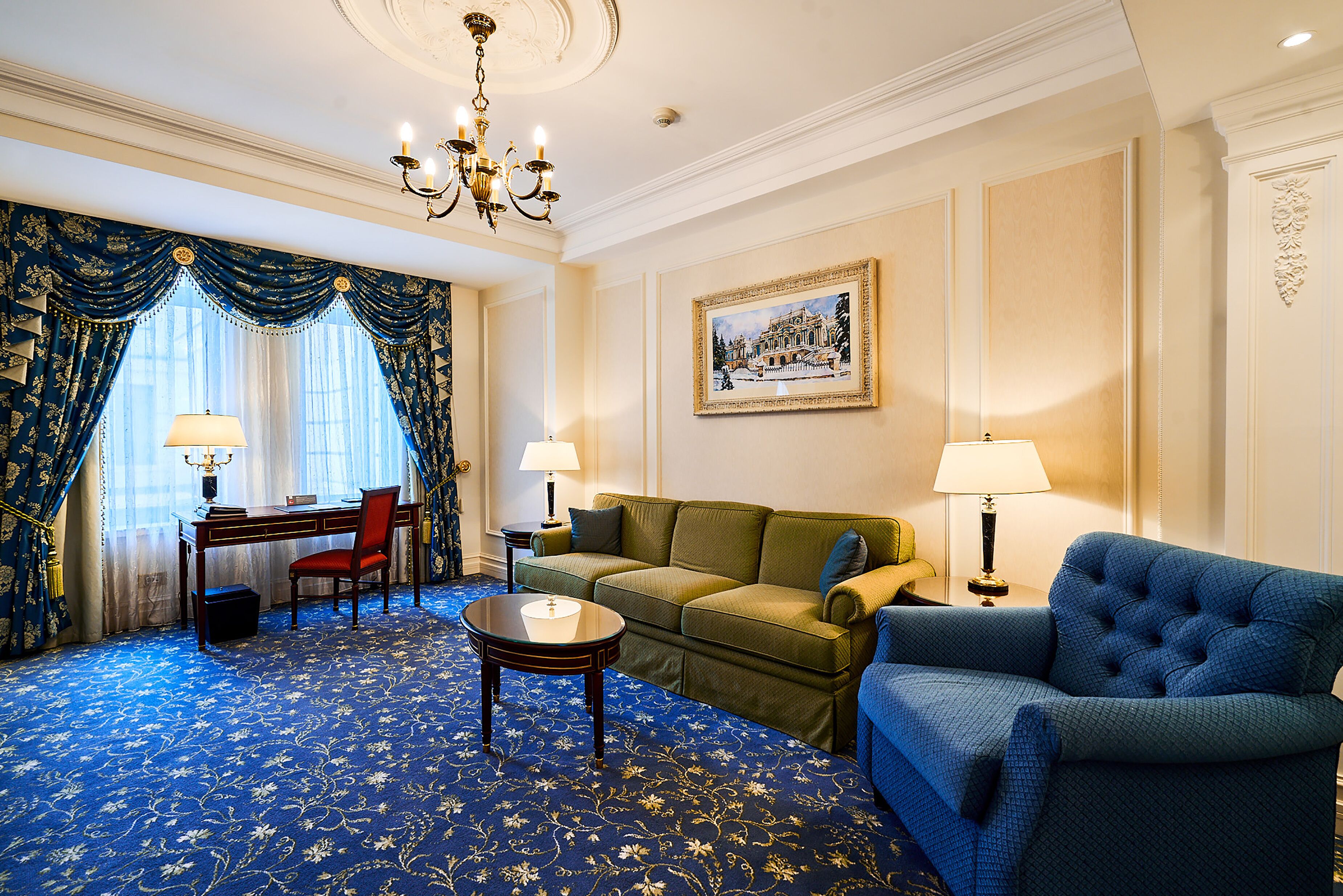 A room with a blue carpet, a green sofa, a blue armchair, a small round table, and a desk with a chair. There is a chandelier on the ceiling and a painting on the wall.