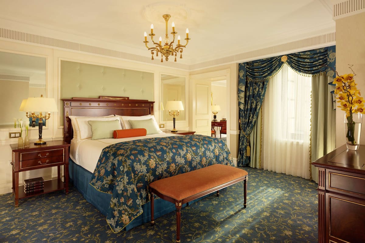 A luxurious bedroom with a large bed, a bench, a chandelier, and ornate curtains.