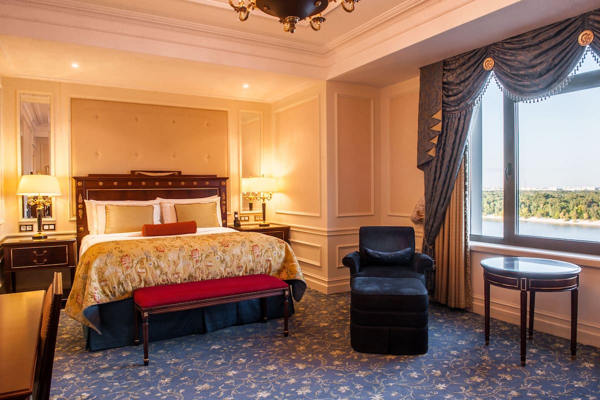 A hotel room with a large bed, a bench, a chair, a small table, and a view of the outdoors.
