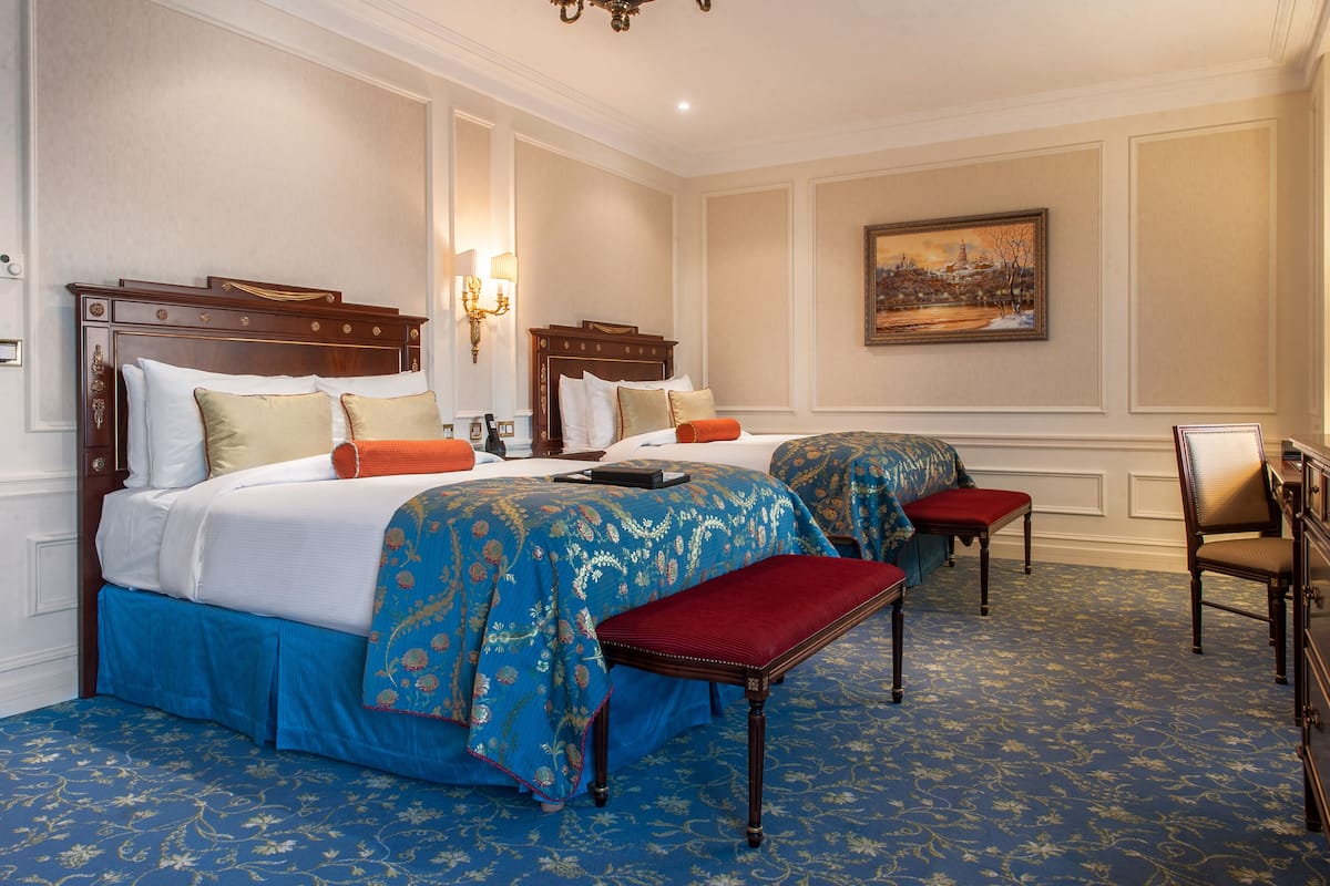 A hotel room with two beds, a red bench, a chair, and a painting on the wall.
