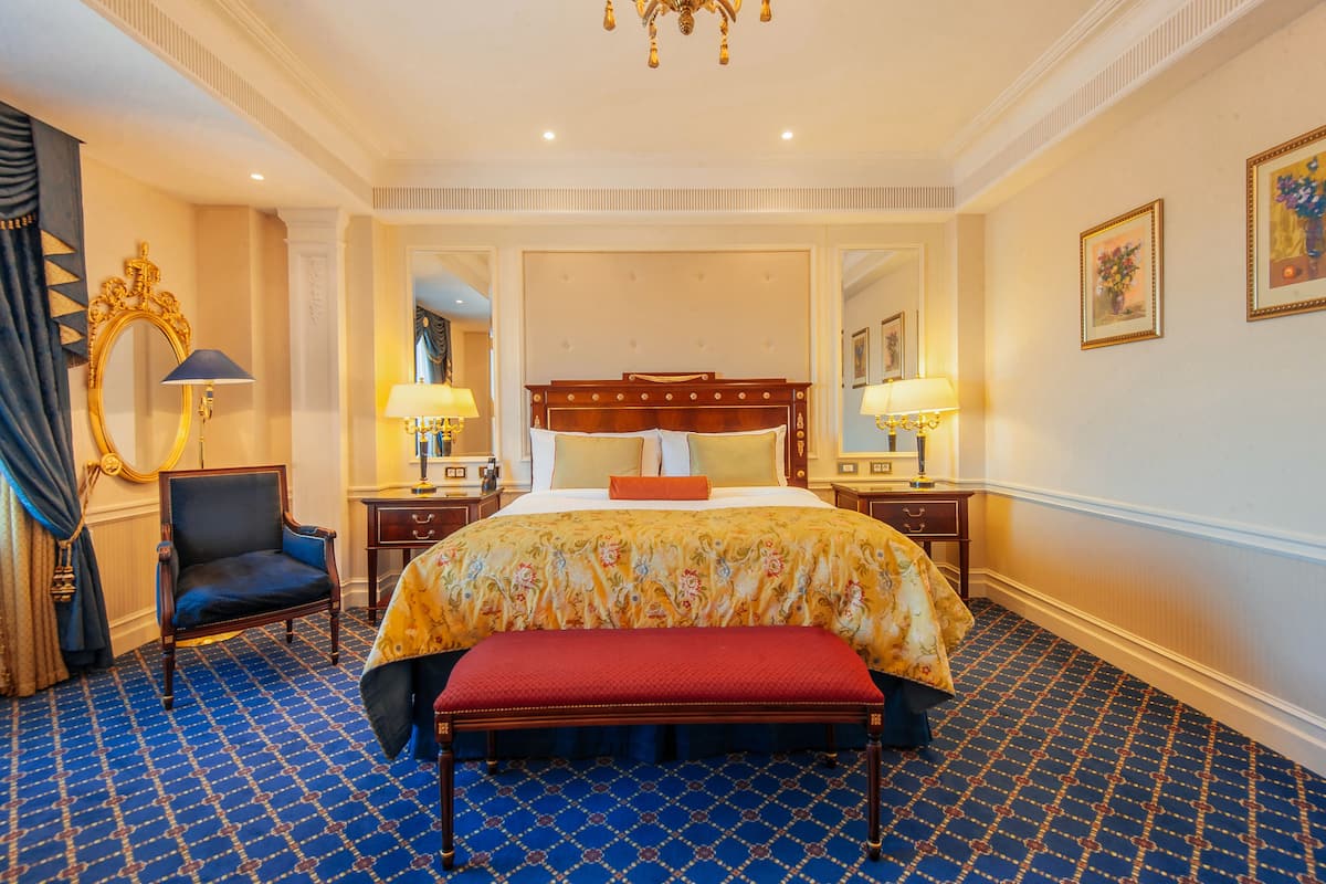 A large bed with a floral bedspread, a red bench, a chair, and two bedside tables with lamps.