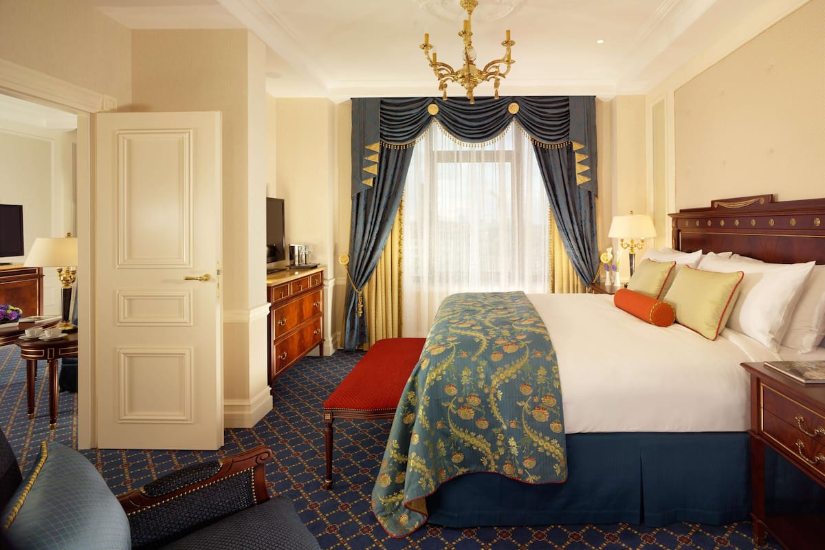 A hotel room with a large bed, a red bench, a wooden dresser, a television, and a chandelier.