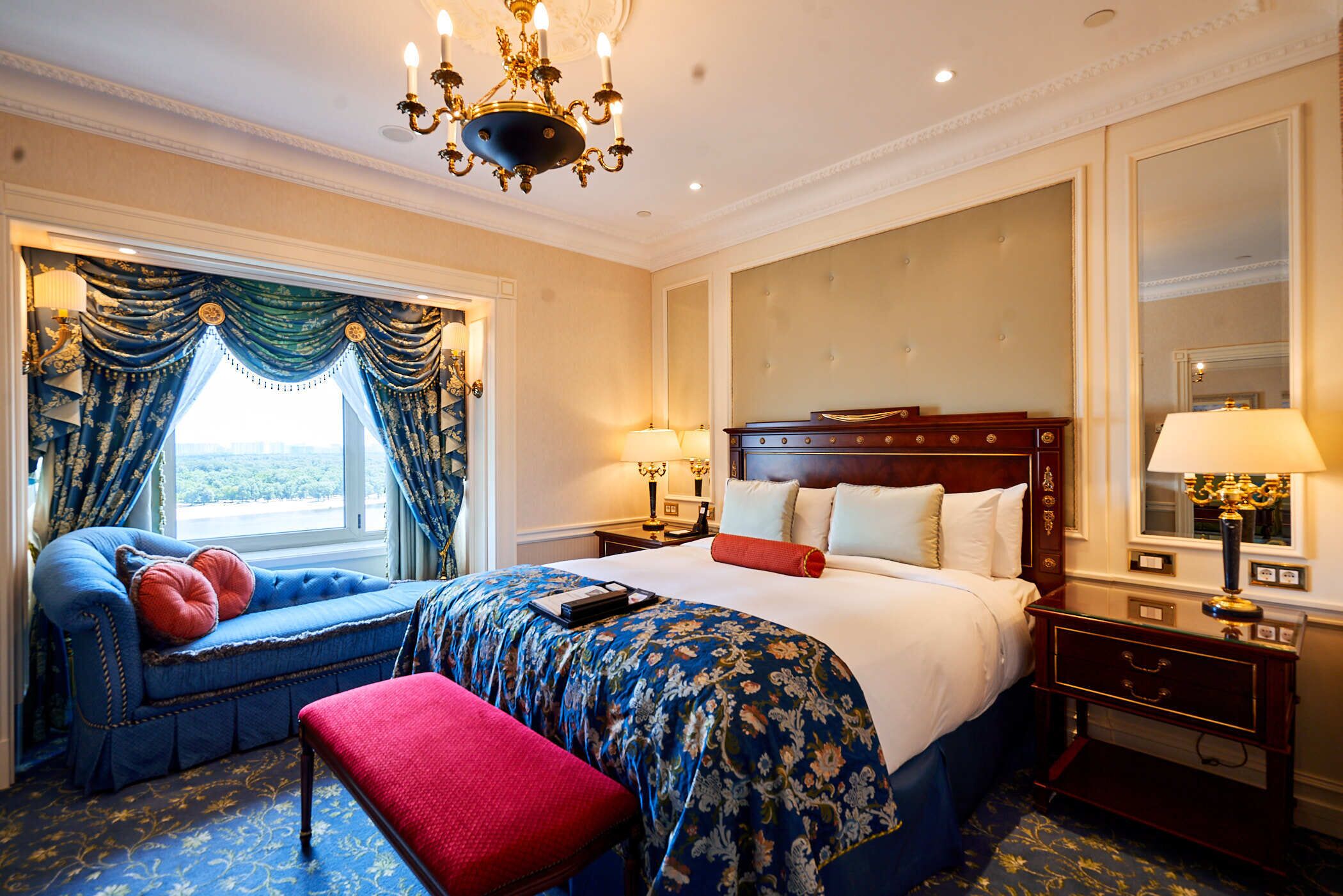 Habitación de hotel con una cama grande, un banco, una araña y vistas al exterior.