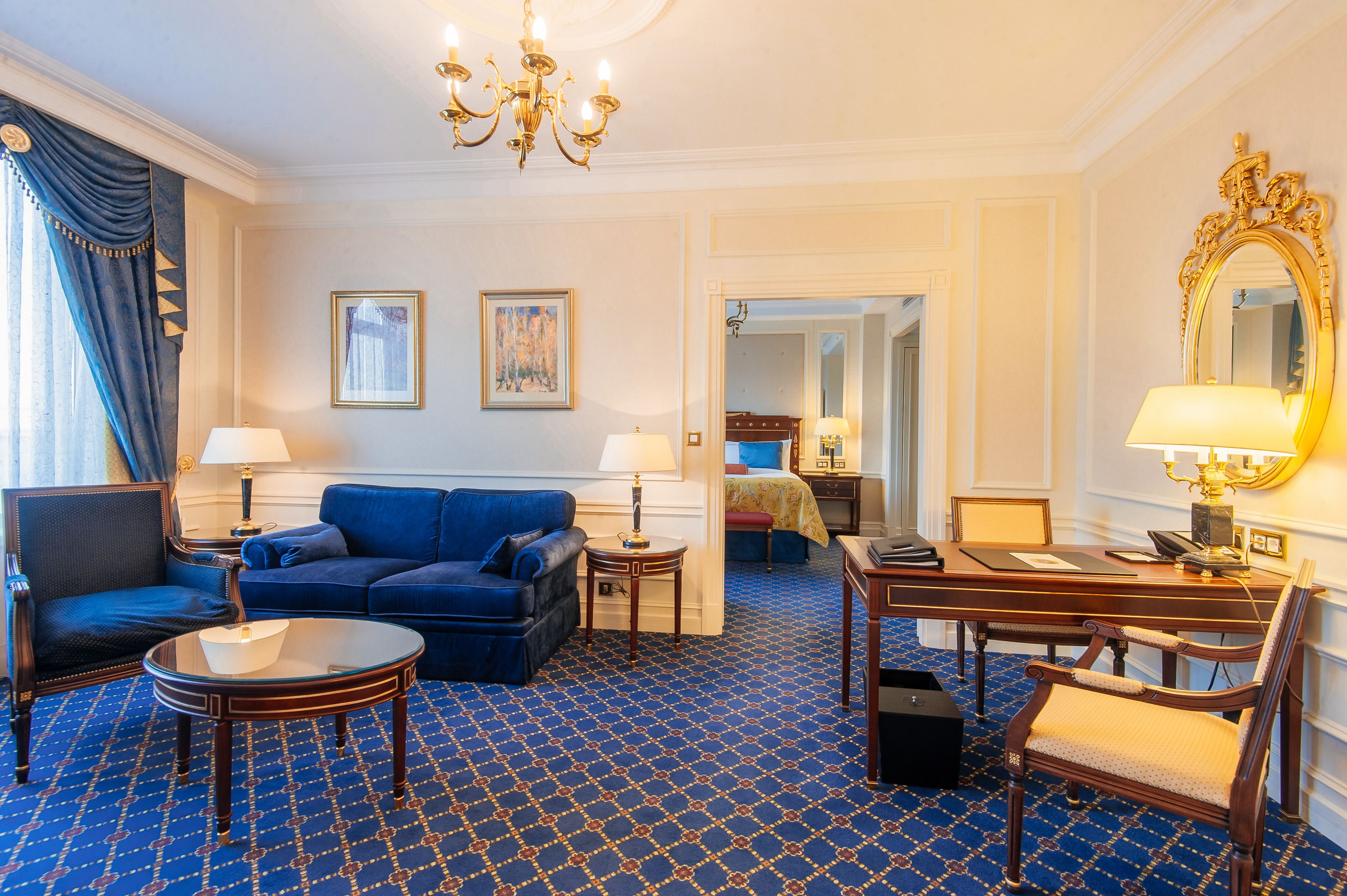 Een hotelkamer met een blauw kamerbreed tapijt, een blauwe bank, een houten bureau, een stoel, een lamp, een spiegel en een bed met een blauw hoofdeinde.
