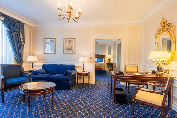 Una habitación de hotel con alfombra azul estampada, un sofá azul, un escritorio de madera, una silla, una lámpara, un espejo y una cama con cabecera azul.