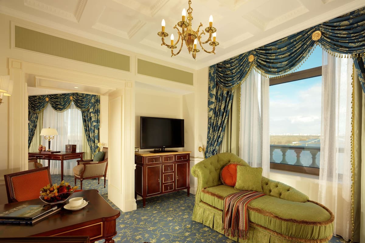 A hotel room with a large window, a green sofa, a TV, and a dining area with a table and chairs.