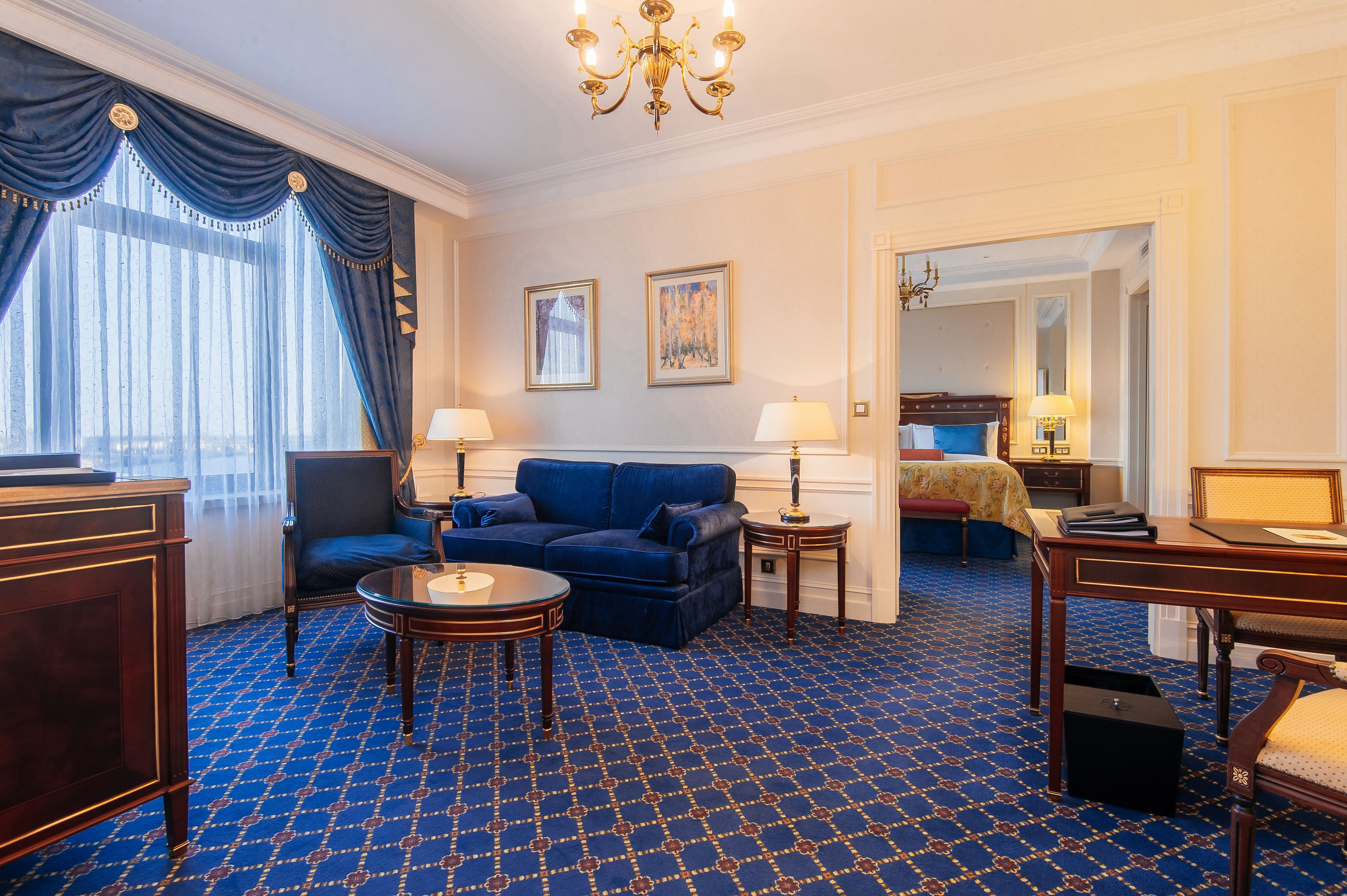 A hotel room with a blue patterned carpet, a blue sofa, two bedside tables with lamps, a wooden desk, and a chair.