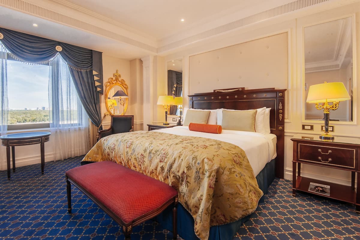 A hotel room with a large bed, a bench, a nightstand, a mirror, and a window with curtains.
