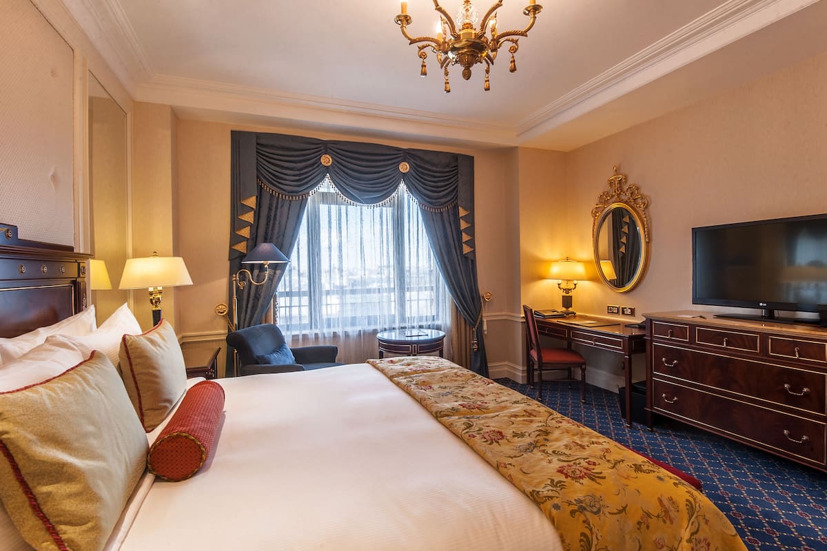 A hotel room with a large bed, a desk with a chair, a television, and a chandelier.