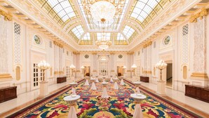 Een statige hotellobby met een hoge plafond, gedetailleerde kroonluchters en een groot bloementapijt.
