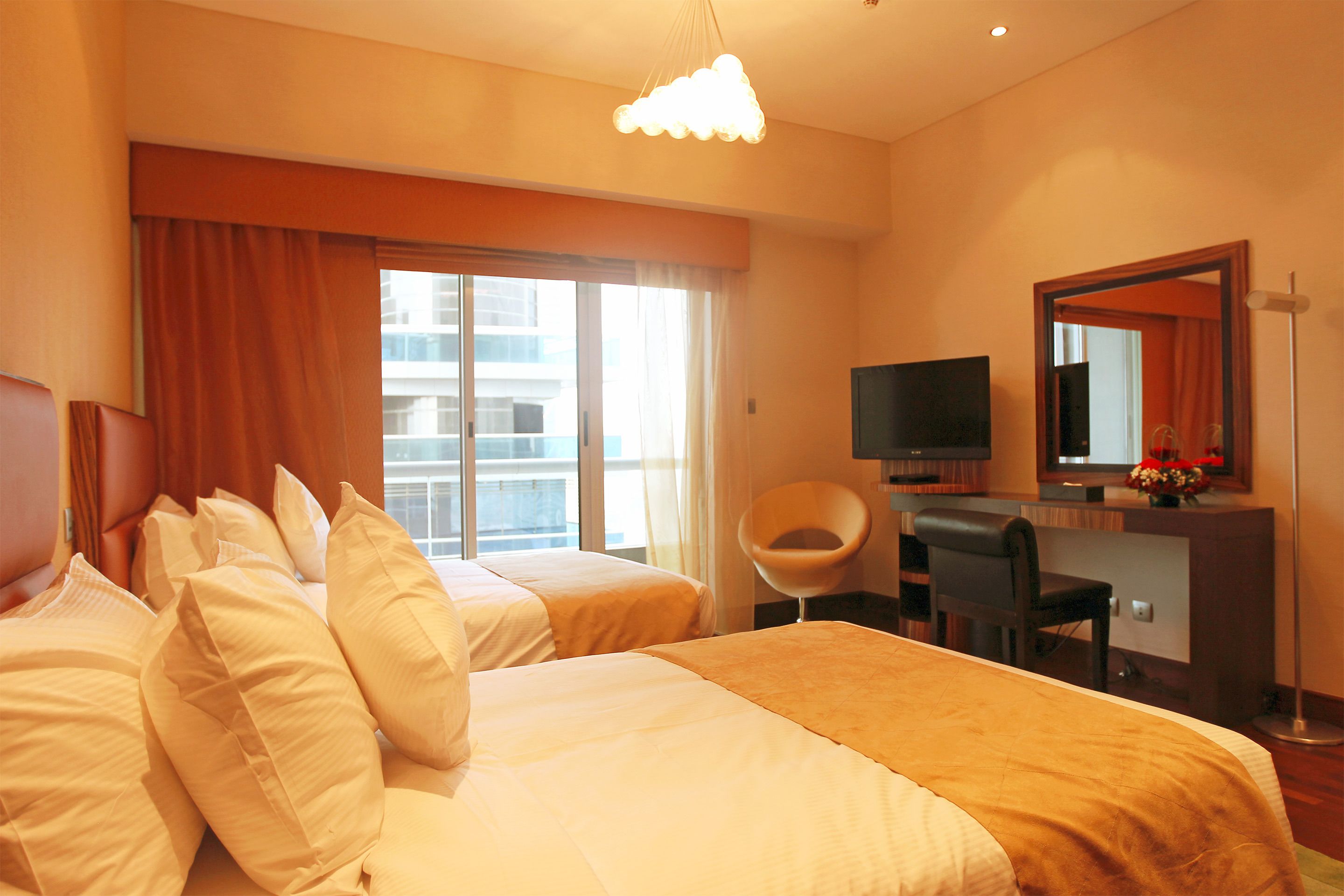 A hotel room with two beds, a desk with a chair, a television, and a large window with curtains.