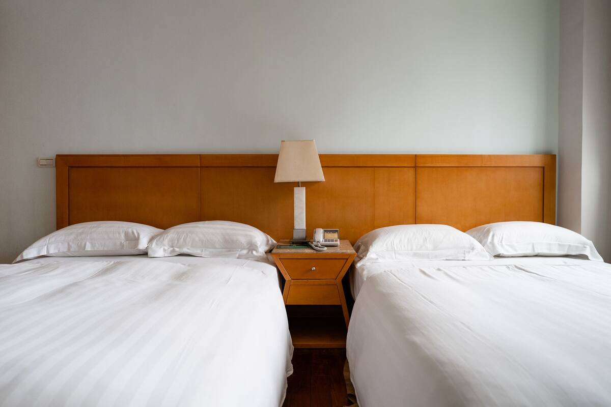 A hotel room with two beds, a wooden headboard, a bedside table with a lamp, and a phone.