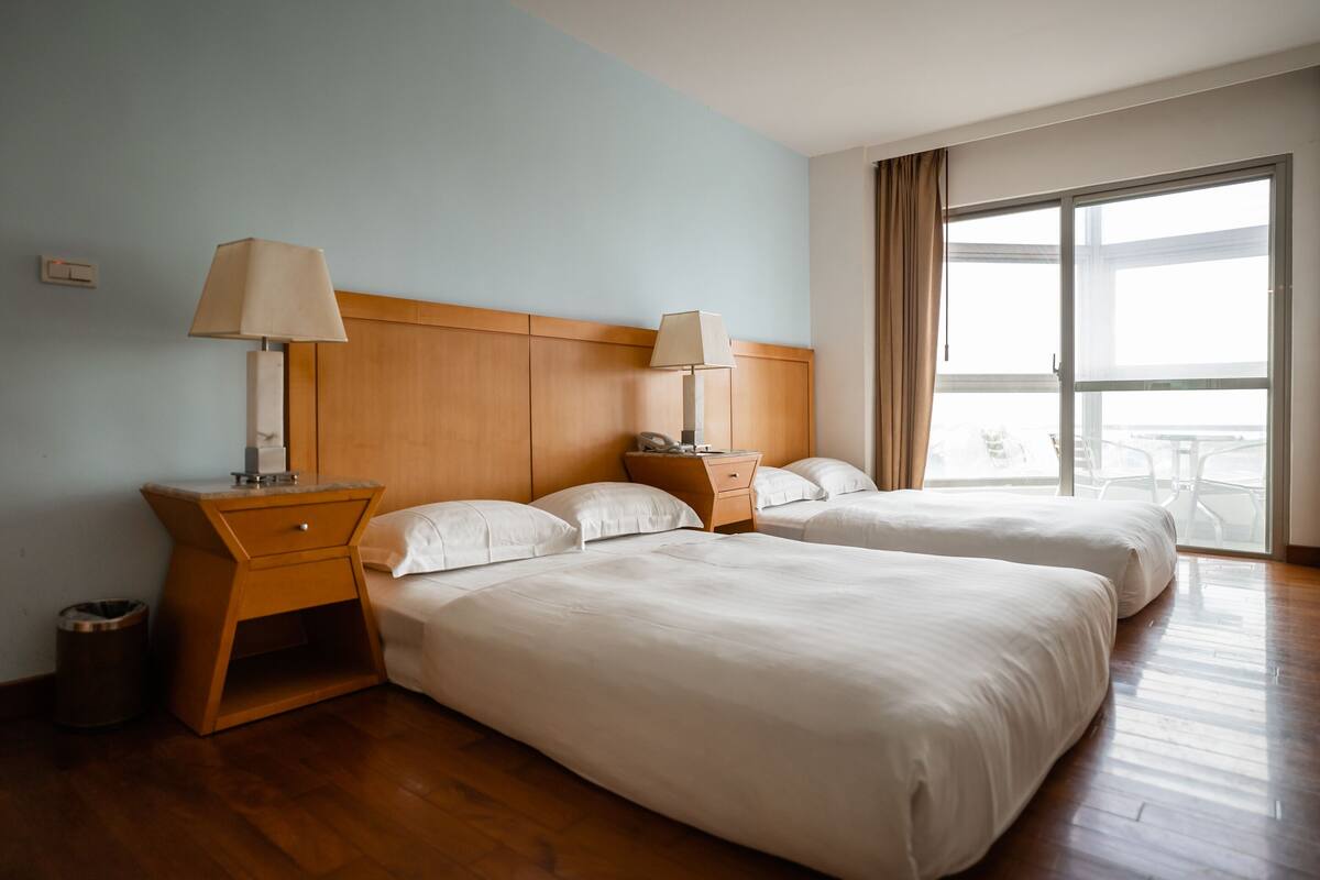 A hotel room with two beds, wooden headboard, bedside tables with lamps, a small trash bin, and a large window with a view of the outside.