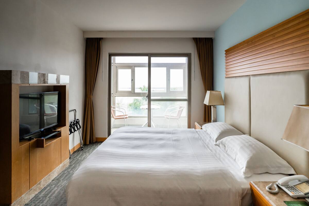 A hotel room with a large bed, a television, a desk with a telephone, and a view through a glass door.