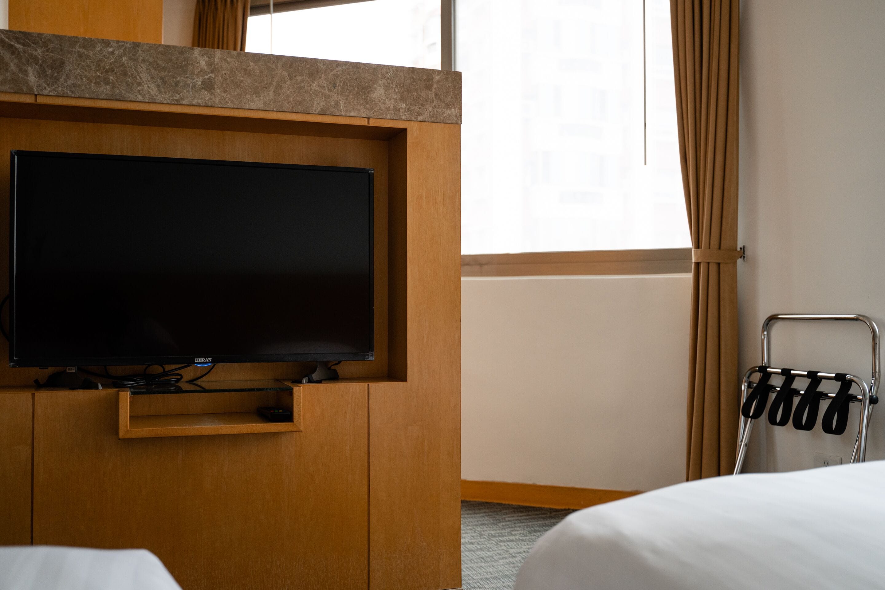 Une chambre d’hôtel dotée d’un téléviseur à écran plat fixé sur un panneau en bois, d’une petite étagère en dessous et d’un porte-serviettes en métal avec des serviettes.