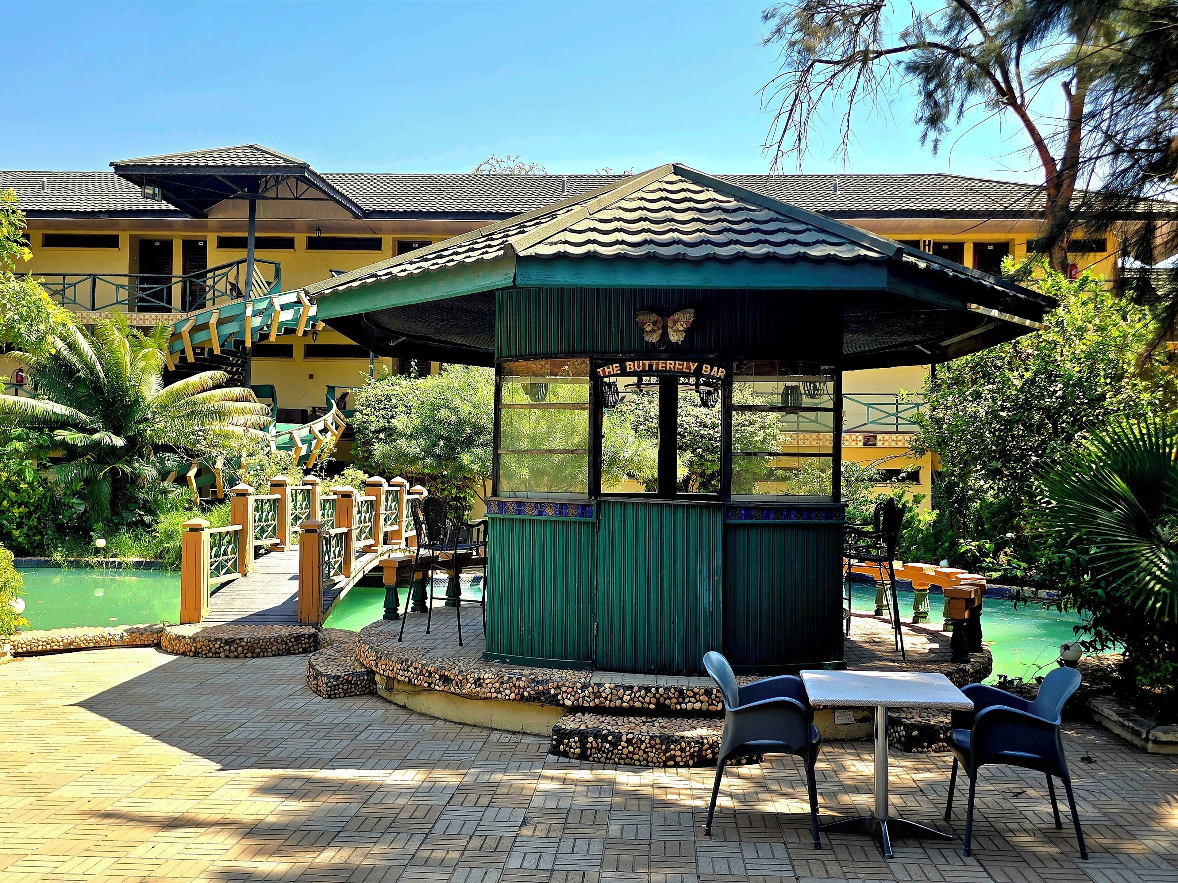 Ein Pavillon mit einem Schild, auf dem 'The Butterfly Bar' steht, umgeben von einem Pool, Stühlen und einer Brücke.
