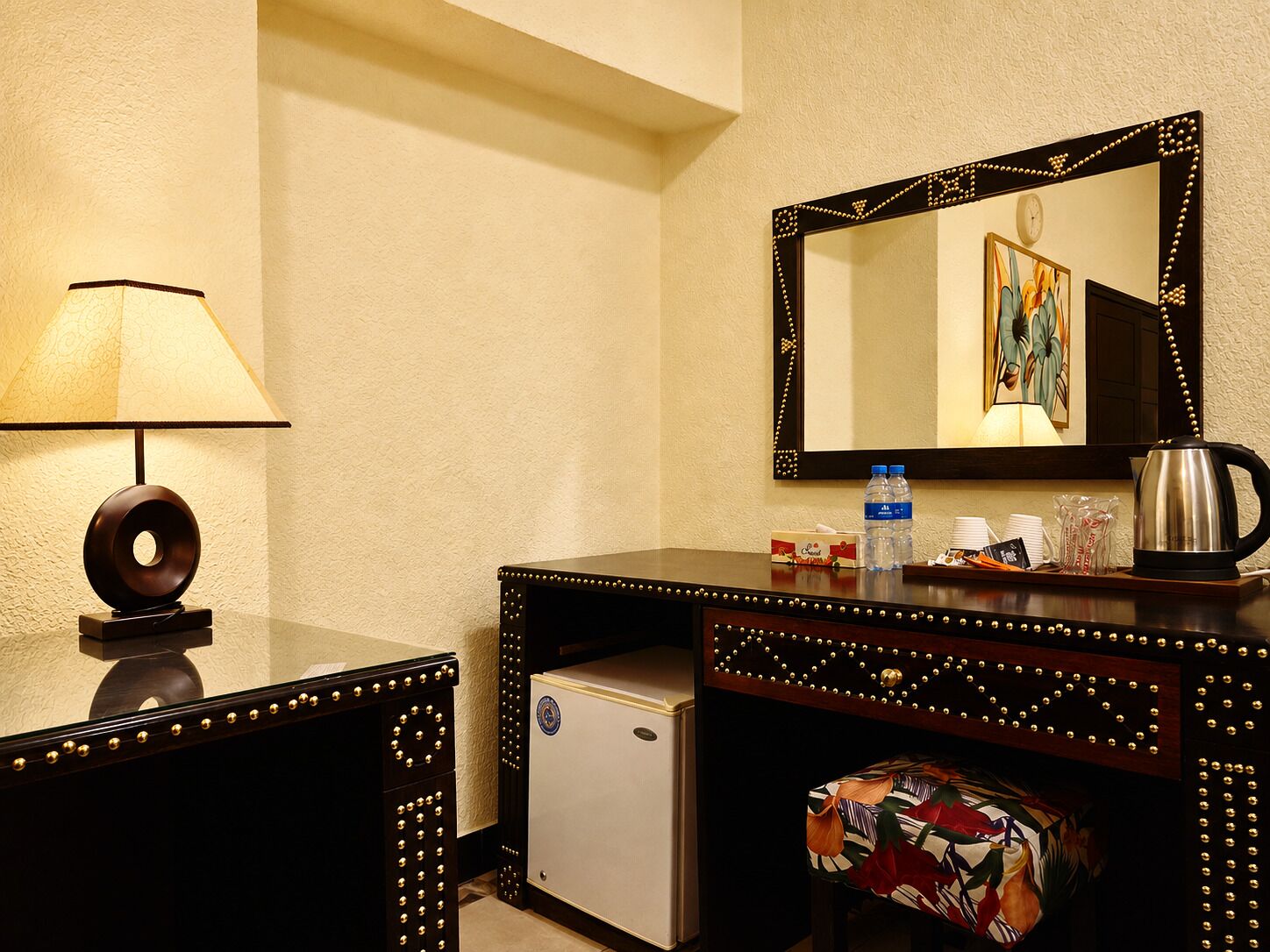A hotel room with a desk, a lamp, a refrigerator, a coffee maker, and a mirror with a decorative frame.