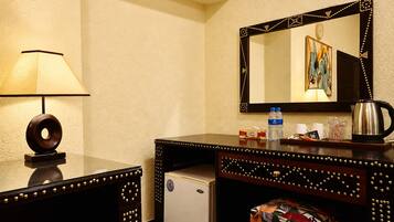 A hotel room with a desk, a lamp, a refrigerator, a coffee maker, and a mirror with a decorative frame.