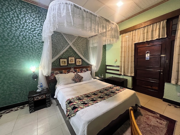 A bedroom with a bed, a nightstand, a bedside table, a wall-mounted shelf, a door, and a window with curtains. - New Dodoma Hotel (Dodoma)