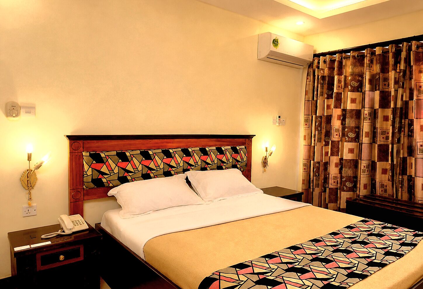 A hotel room with a bed, bedside lamps, a telephone, an air conditioning unit, and patterned curtains.