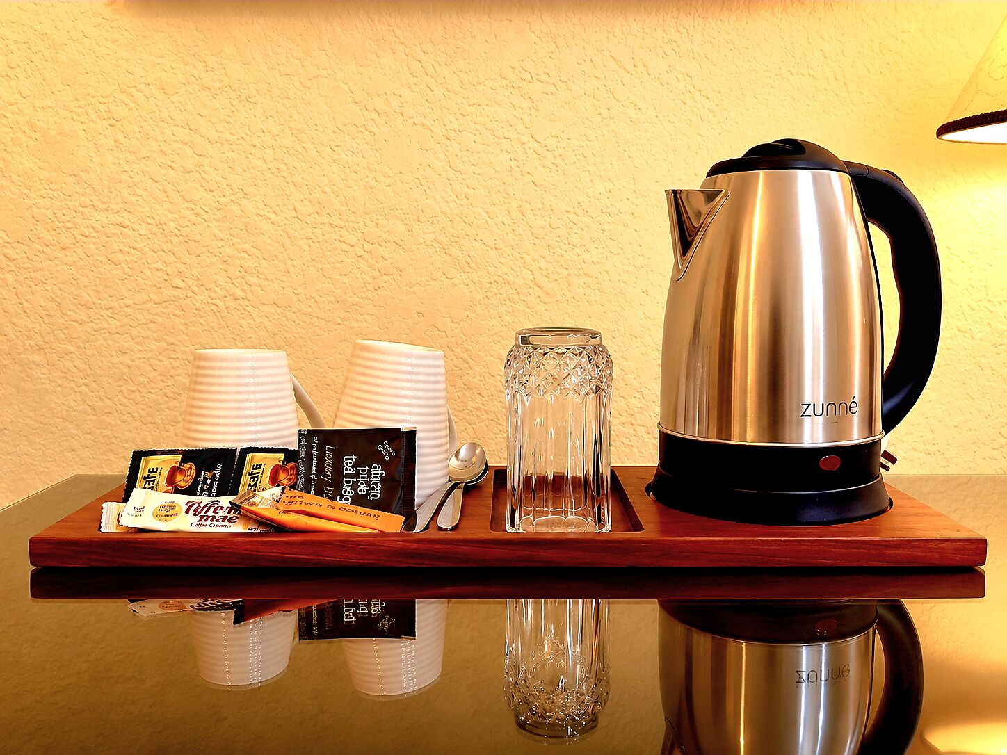 A tray with a stainless steel kettle, two white cups, a glass, and coffee packets on a reflective surface.