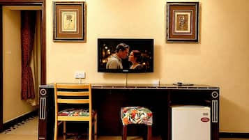 Una habitación con televisión montada en la pared, un refrigerador pequeño, una silla de madera y un banco con cojín floral.