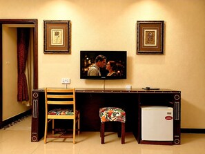 Una habitación con televisión montada en la pared, un refrigerador pequeño, una silla de madera y un banco con cojín floral.