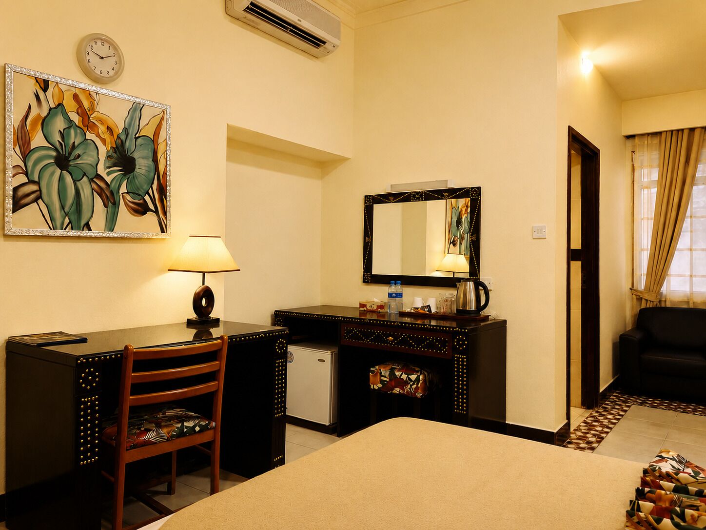 A hotel room with a bed, desk, chair, and a small refrigerator. There is a painting on the wall, a clock, and a mirror.