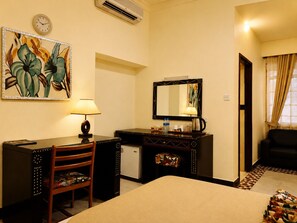A hotel room with a bed, desk, chair, and a small refrigerator. There is a painting on the wall, a clock, and a mirror. - New Dodoma Hotel (Dodoma)