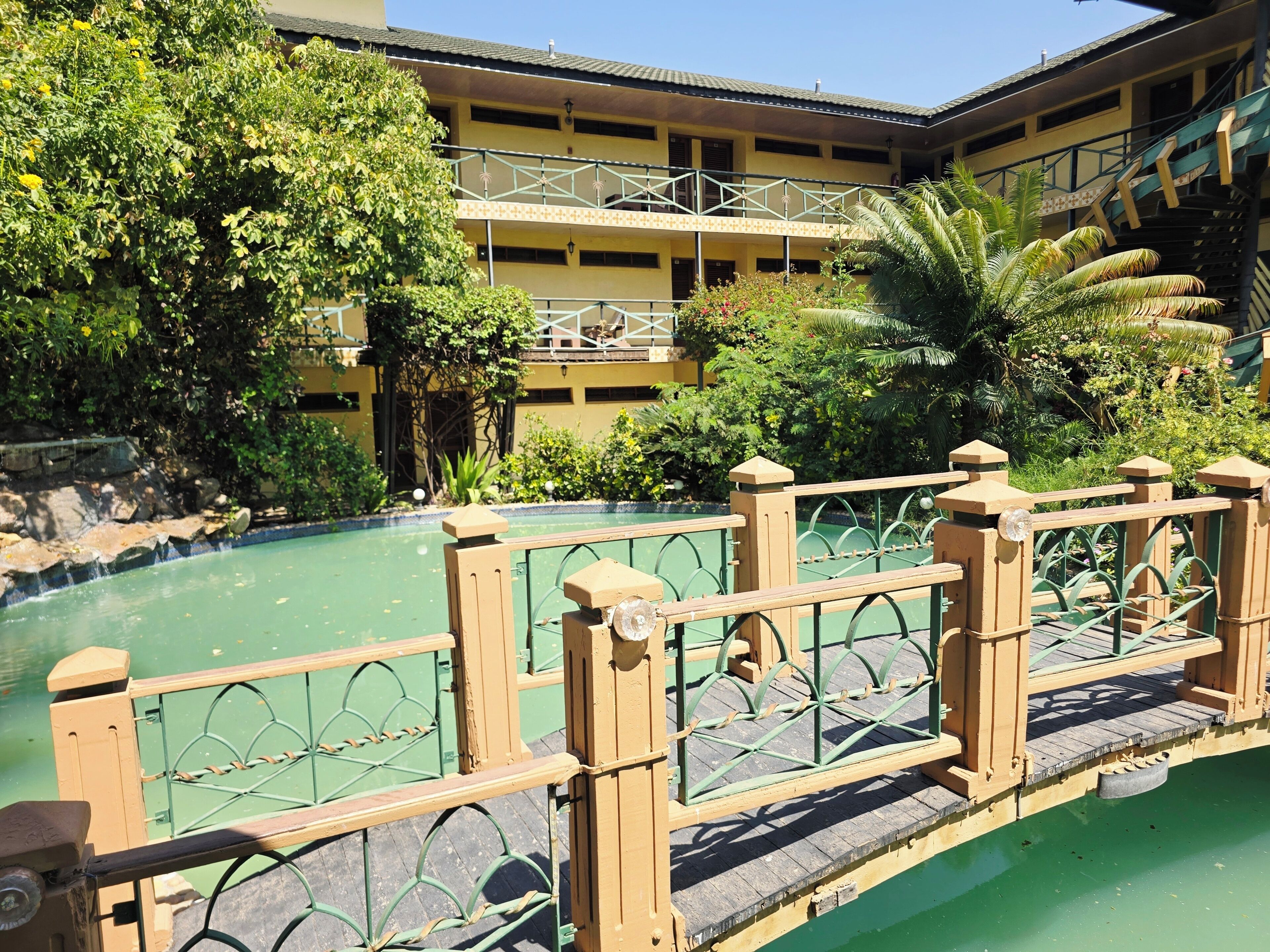Un área de piscina con valla de madera, rodeada de abundante vegetación y un edificio de varios pisos con balcones.