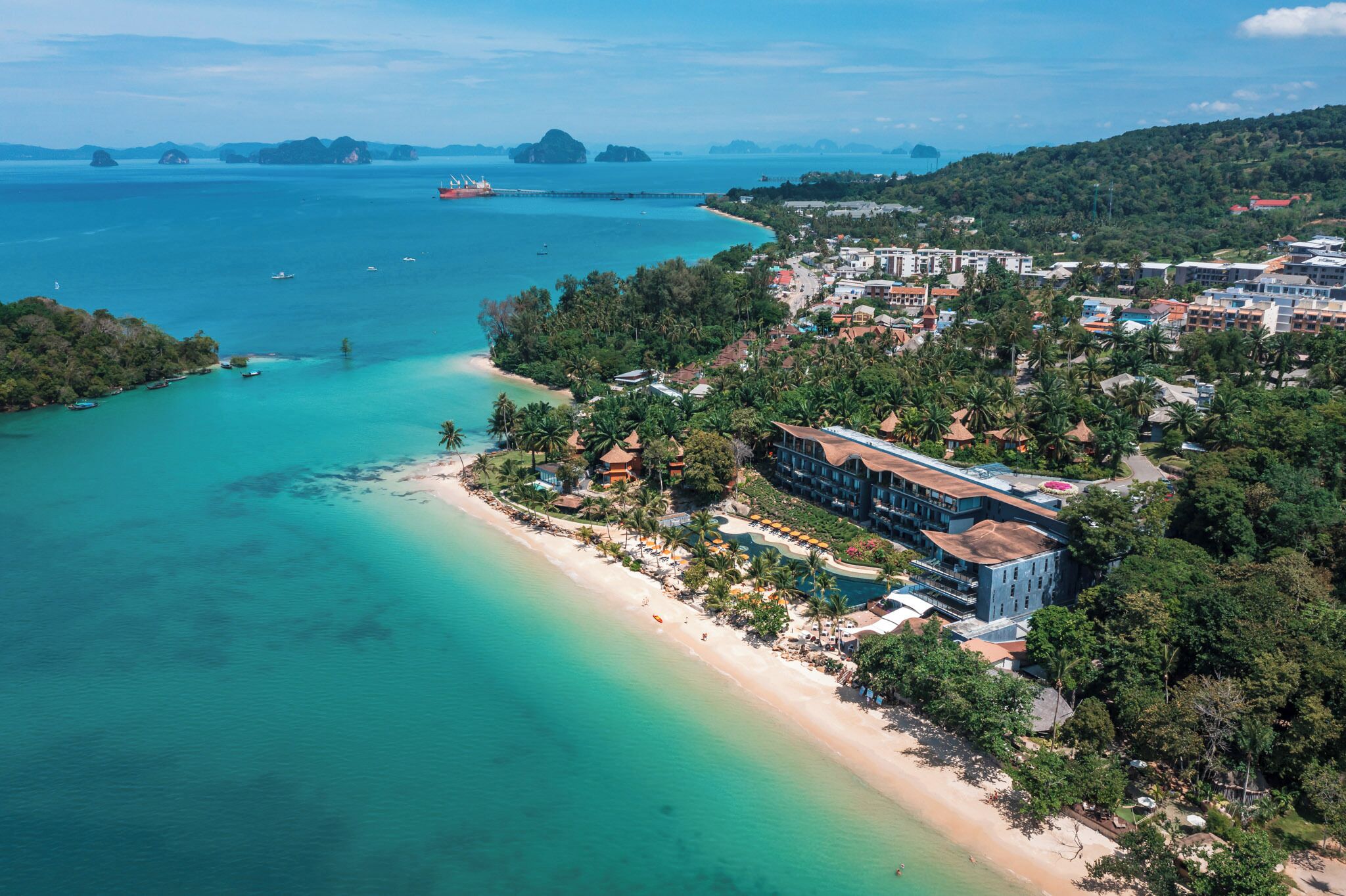 An aerial view of a coastal resort with a beachfront, a large building with multiple floors, and a clear view of the surrounding water and islands.
