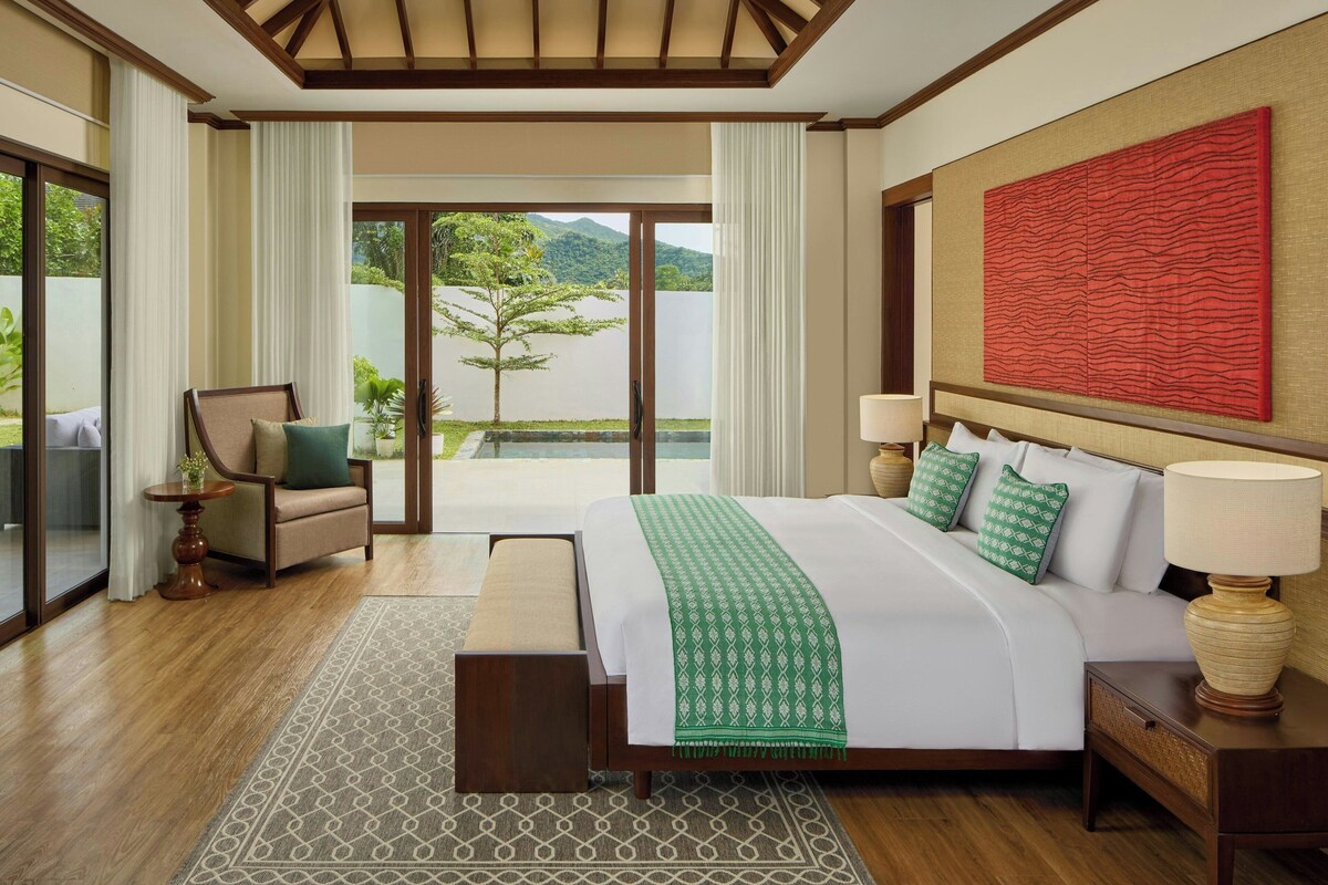 A hotel room with a large bed, a chair, a table, and a view of a garden and mountains.