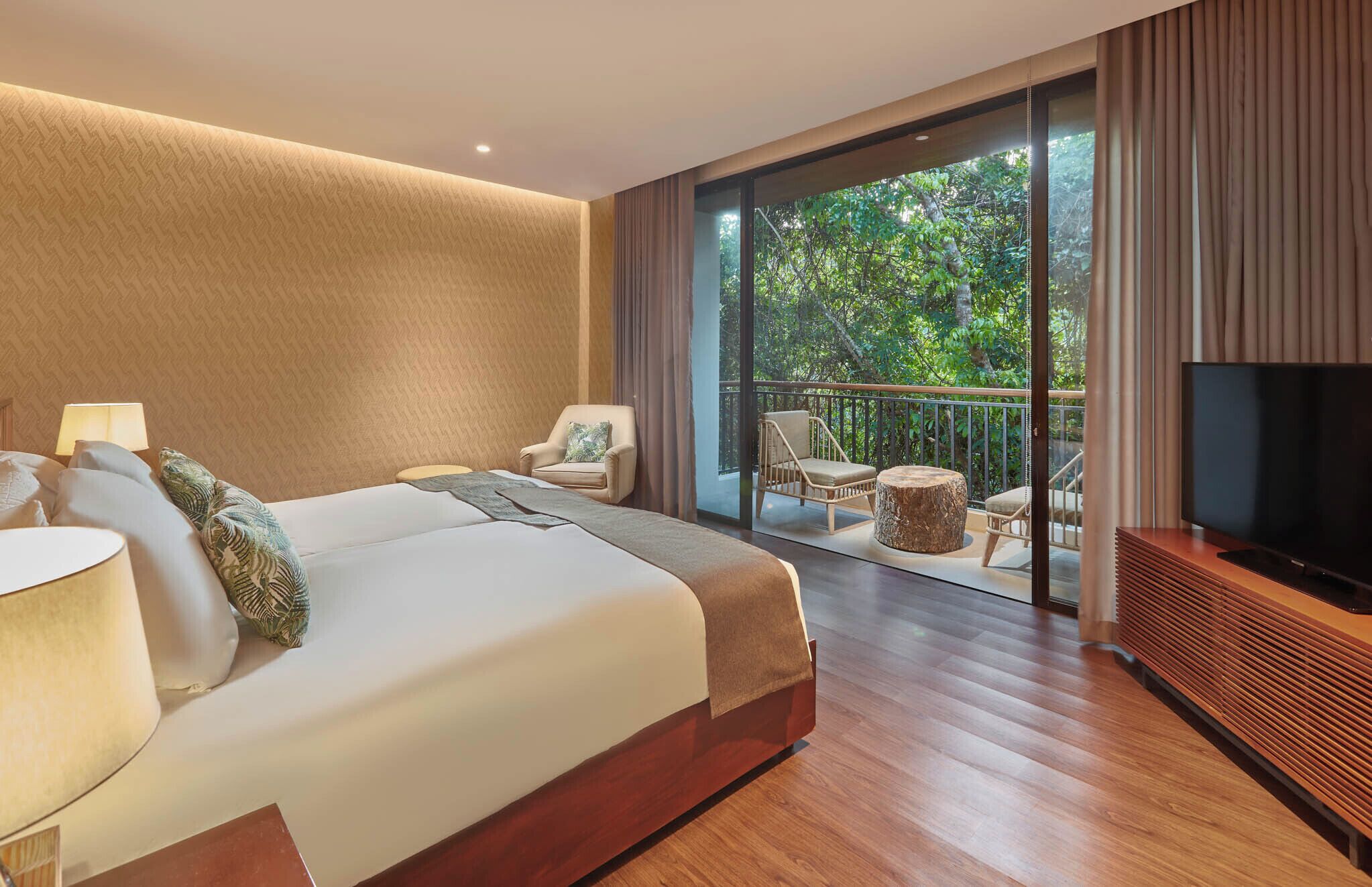 Een hotelkamer met een groot bed, een televisie, een balkon met tuinmeubilair en uitzicht op groen.