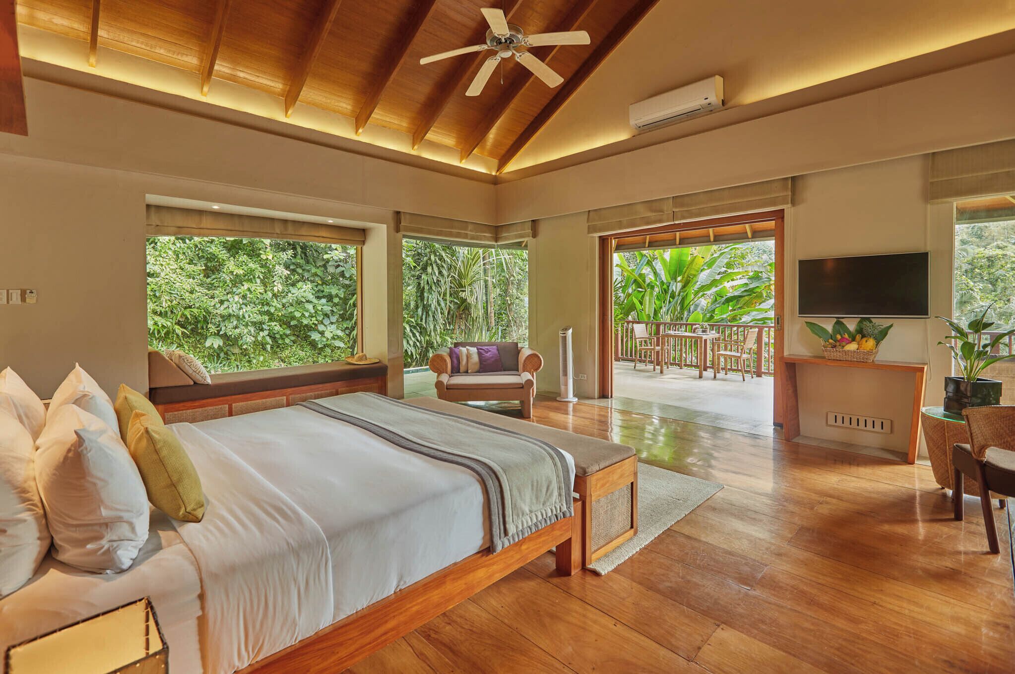 A spacious bedroom with a large bed, wooden ceiling, and a view of lush greenery outside.