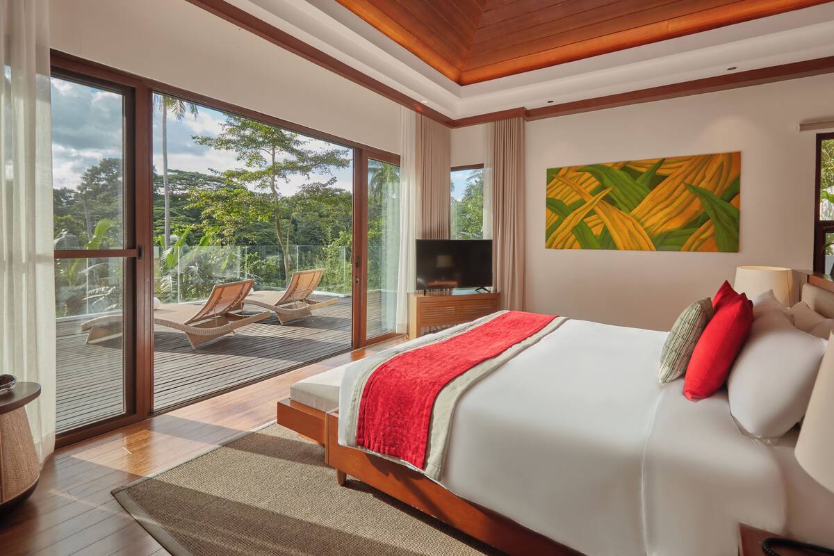 A bedroom with a bed, a TV, a balcony with chairs, and a view of greenery.