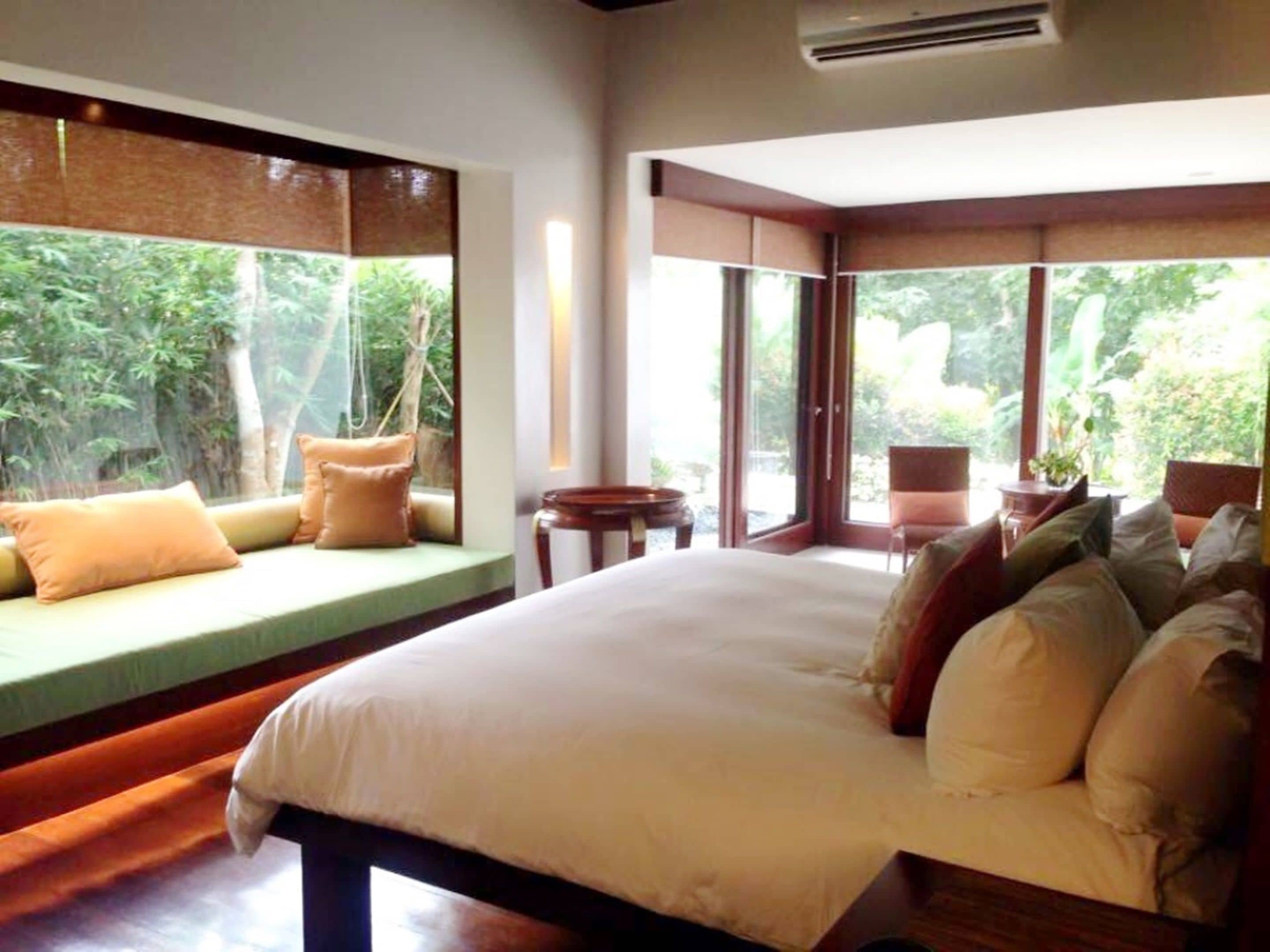 A large bed with multiple pillows, a small table, and a chair in a room with large windows overlooking greenery.
