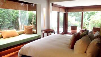 A large bed with multiple pillows, a small table, and a chair in a room with large windows overlooking greenery.
