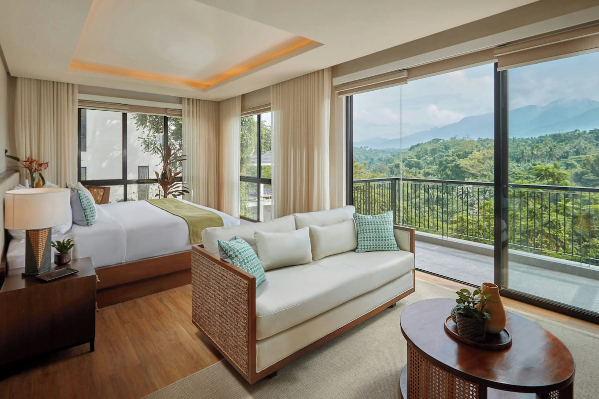 A hotel room with a bed, a sofa, a nightstand, a lamp, a vase, and a view of a lush landscape.