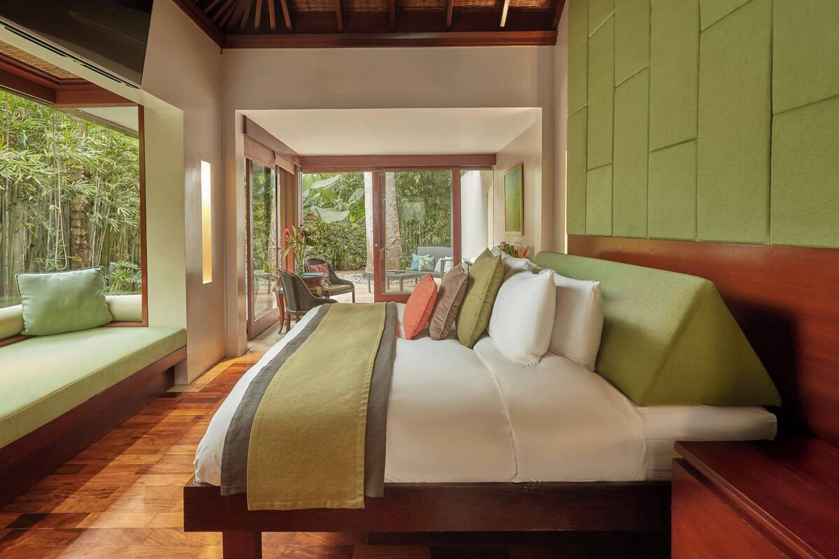 A spacious bedroom with a large bed, a seating area with cushions, and a view of greenery outside.