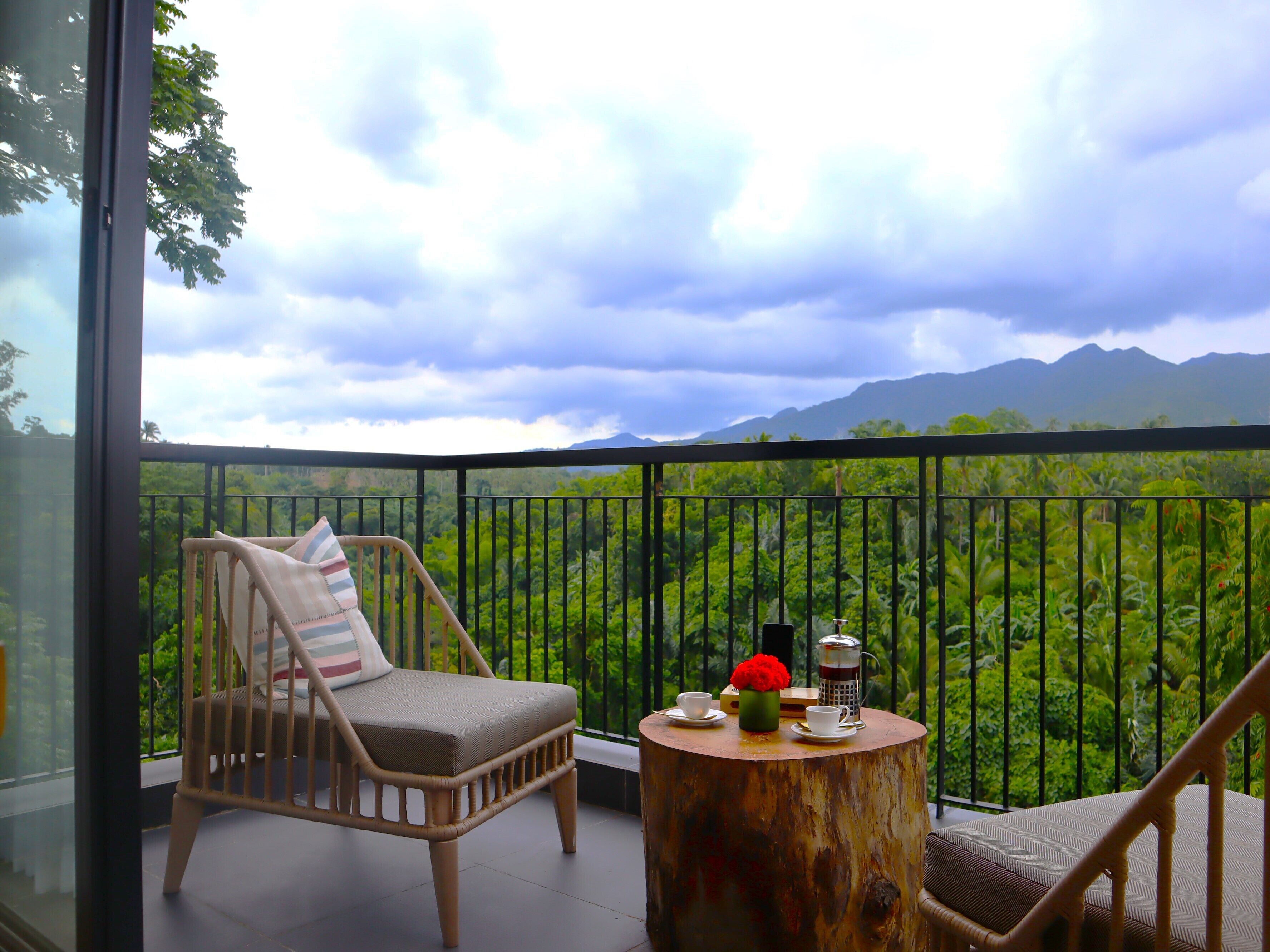 A balcony with a wicker chair, a small round wooden table, and a view of a lush green landscape with mountains in the distance.