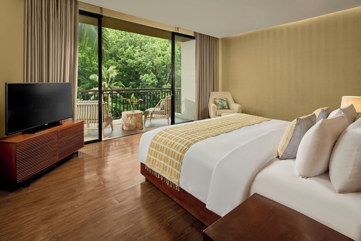 Una habitación de hotel con una cama grande, un televisor de pantalla plana sobre un mueble de madera, un balcón con vistas a zonas verdes y una silla.