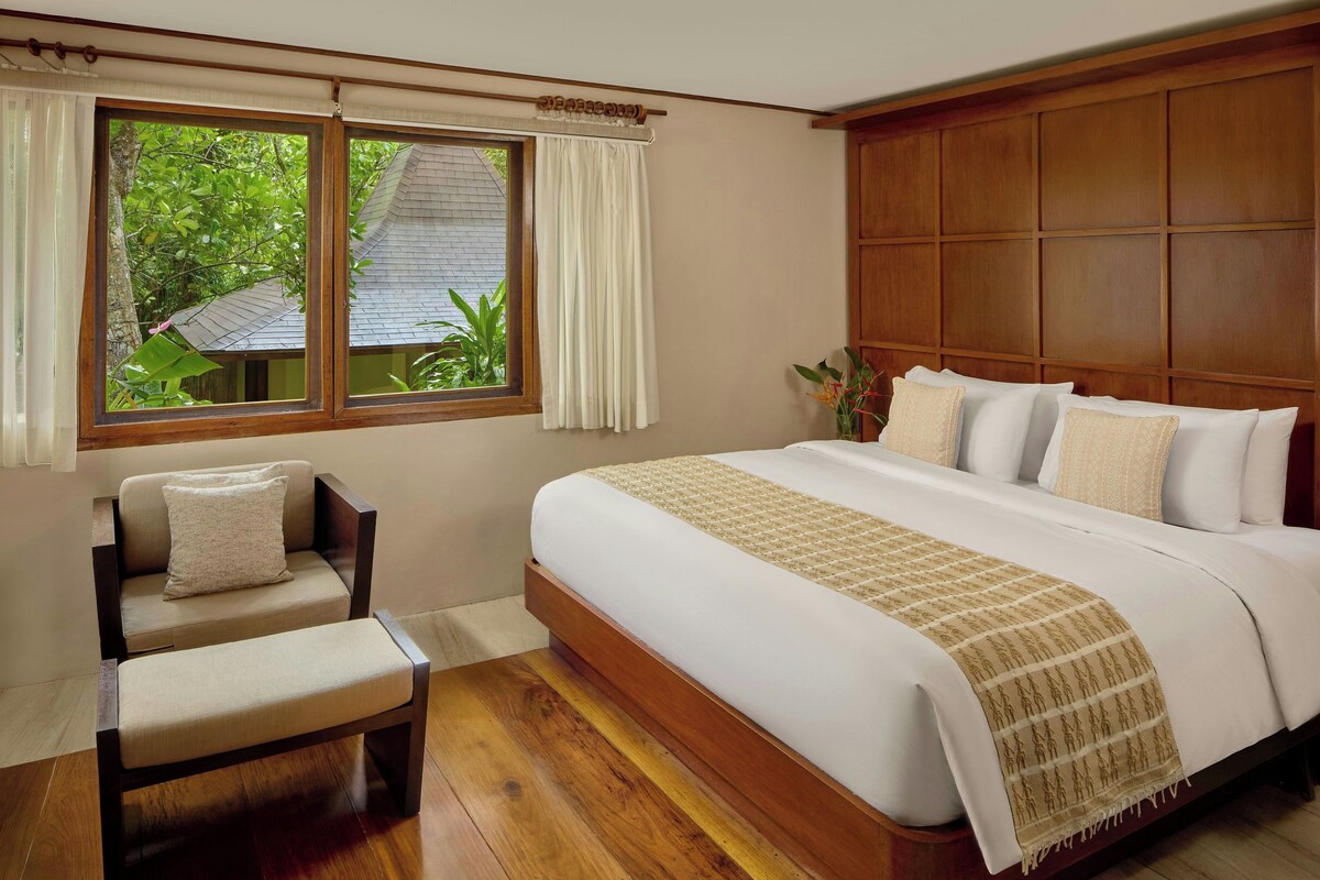 Un dormitorio con una cama grande, cabecera de madera, ventana con cortinas, una silla y vista a zonas verdes.