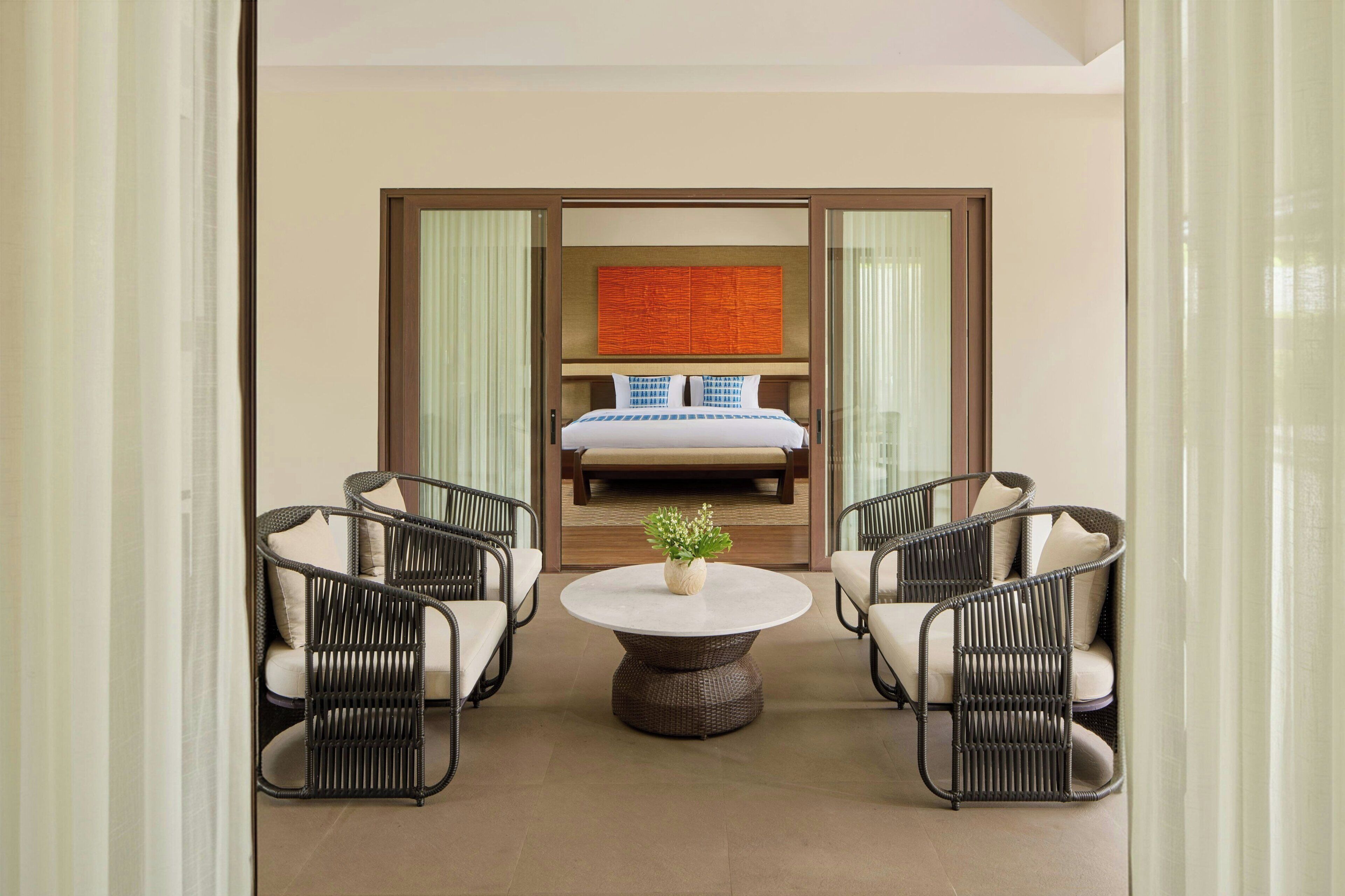A hotel room with a bed, two wicker chairs, a round table, and a decorative plant.