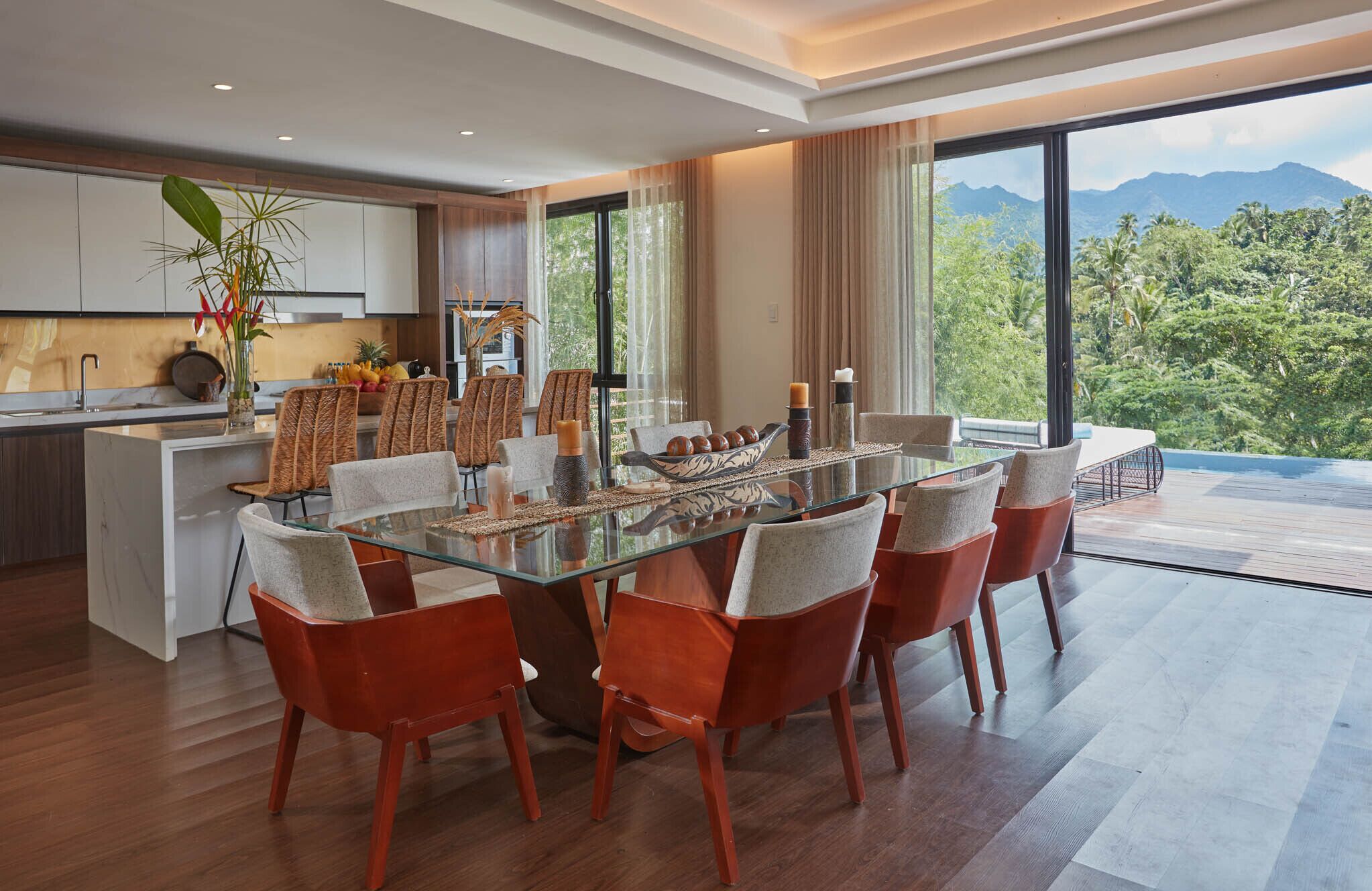 A modern dining area with a glass table, red chairs, and a view of lush greenery and mountains.