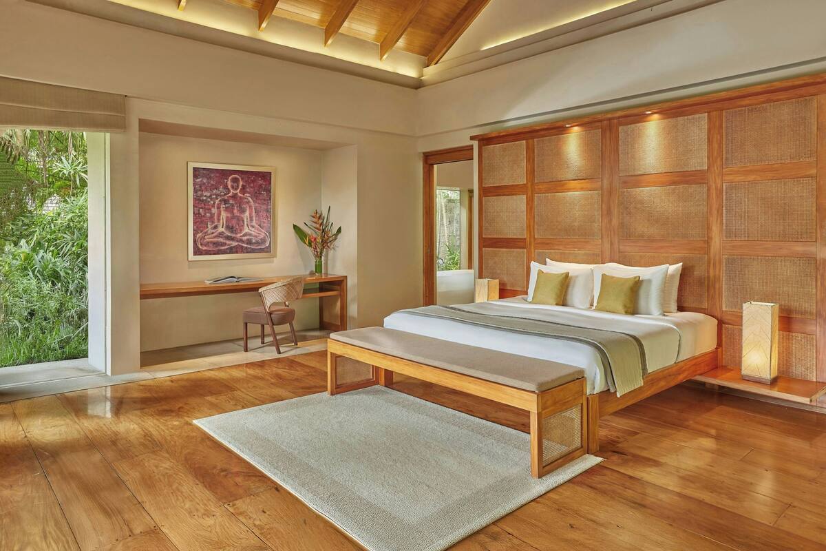 A modern bedroom with a wooden floor, a large bed with pillows, a desk with a chair, and a large window with a view of greenery.