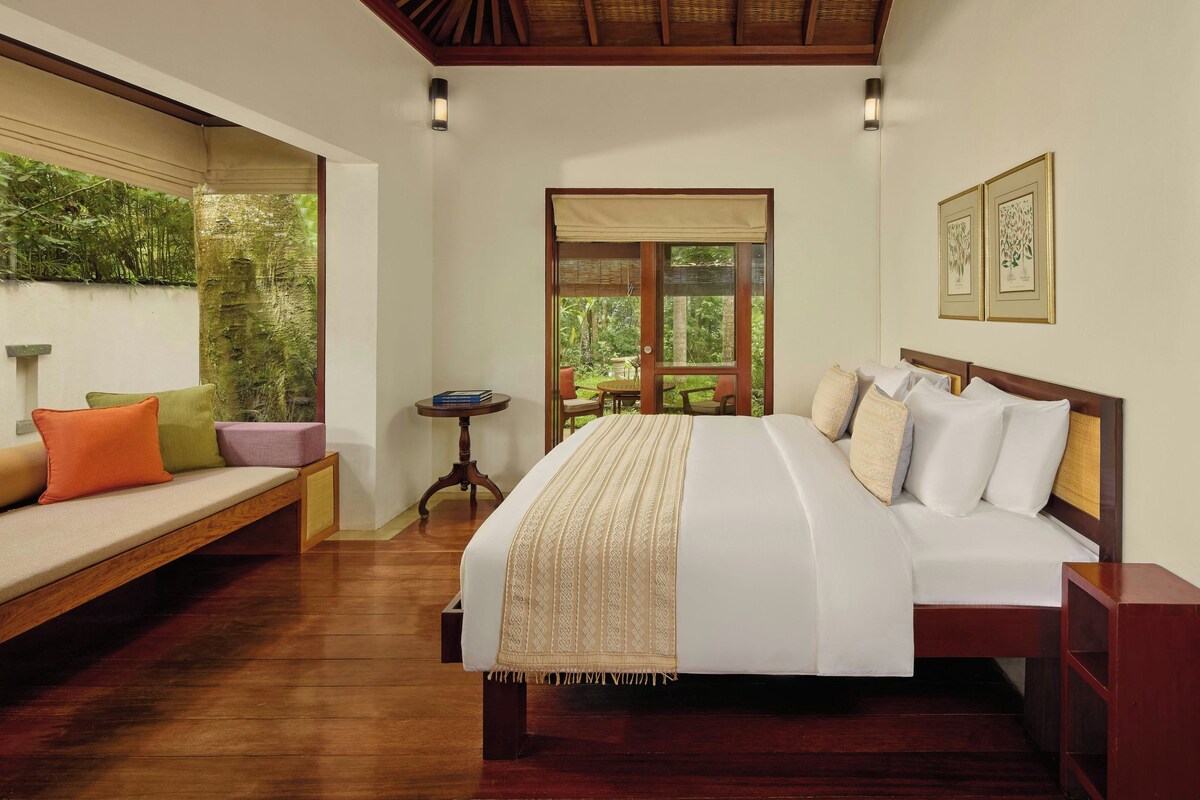 A bedroom with a large bed, a bench with cushions, a small table, and a view of greenery outside.
