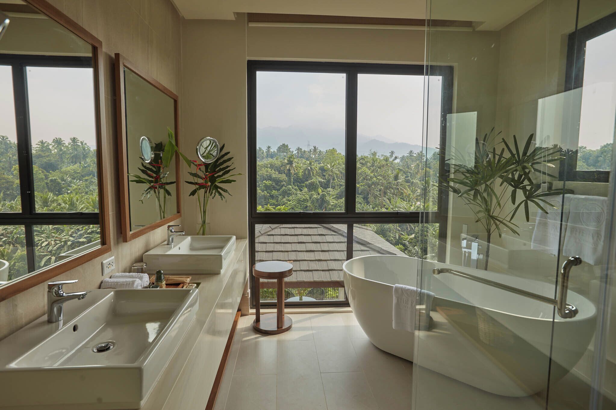Un baño moderno con dos lavamanos, una bañera grande y vistas a un paisaje exuberante a través de amplios ventanales.