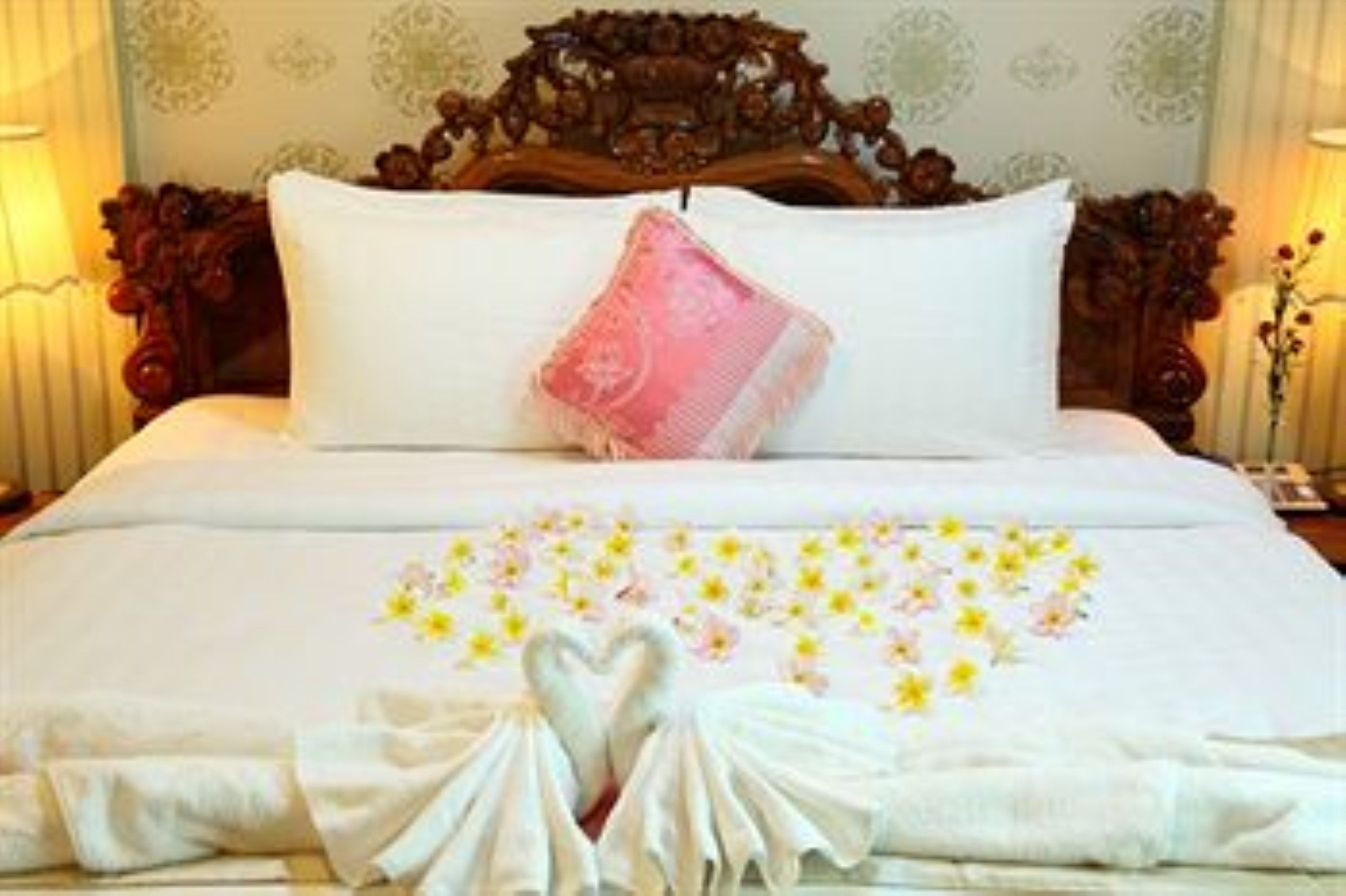 Una cama impecable con un arreglo floral en forma de corazón, una almohada rosa y una toalla blanca a un lado.