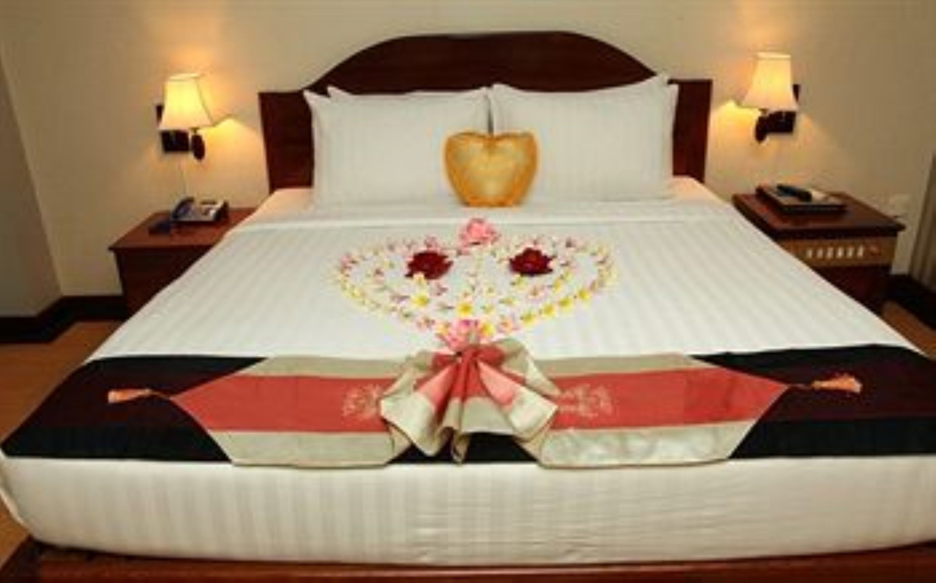 A neatly made bed with a heart-shaped decoration on the bedspread, two bedside lamps, and a wooden headboard.