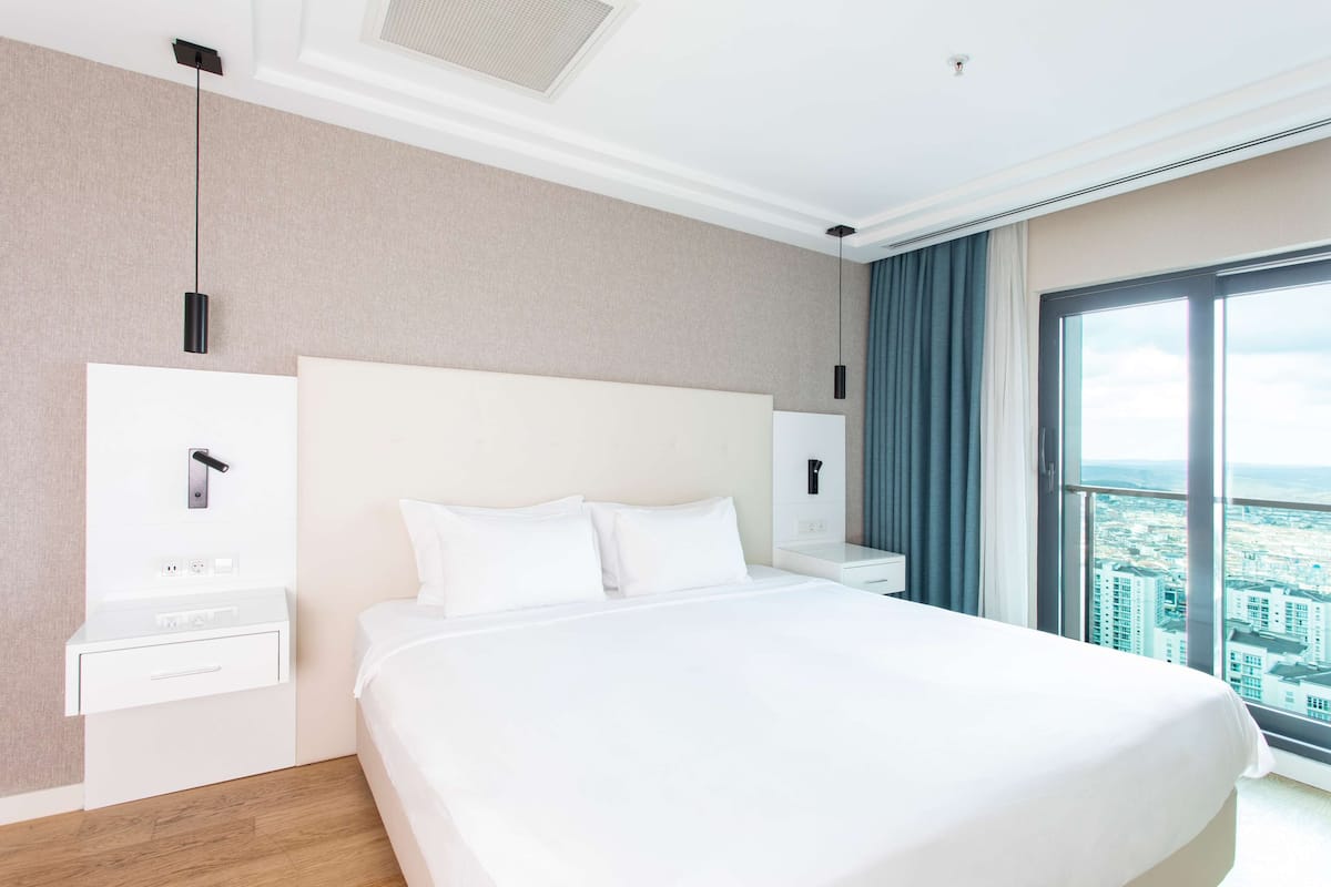 Ein modernes Hotelzimmer mit einem großen Bett, Blick auf die Stadt, einem weißen Nachttisch, einem Wandtelefon und einer Deckenleuchte.